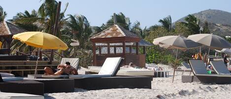 A beach with lounge chairs and umbrellas, a small wooden structure, and palm trees.