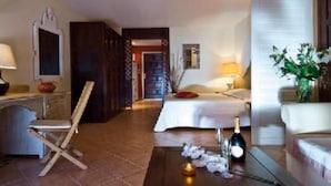 Une chambre d’hôtel avec un lit, un bureau avec une chaise, une petite table avec une bouteille et des fleurs, et une porte donnant sur une autre pièce.