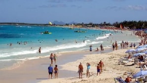 Une plage bondée où l’on voit des gens se promener et nager, et un parachute ascensionnel en arrière-plan.