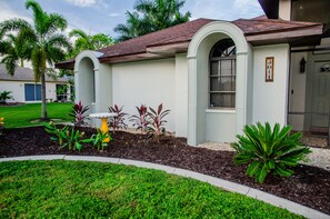 A house with a red roof, white walls, and a small garden with various plants. - Coconut Cove / Scenic Waterfront Escape with Private Heated Pool & Spa (Cape Coral)
