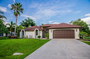 Ein bungalow mit brauner Garagentür, einer kleinen angebauten Garage und einem Vorgarten mit einer Palme und verschiedenen Pflanzen.