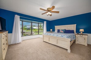 A bedroom with a large bed, a ceiling fan, a nightstand, a dresser, and a window with a view of the outdoors. - Coconut Cove / Scenic Waterfront Escape with Private Heated Pool & Spa (Cape Coral)