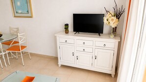 Un salon avec une table basse en verre, un meuble blanc, un téléviseur à écran plat et des fauteuils orange.