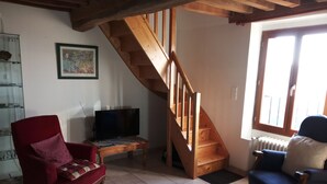 Una sala de estar con una escalera de madera, un sillón rojo, un sillón azul, un televisor sobre una mesa y un cuadro enmarcado en la pared.