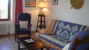 Un salón acogedor con un sillón azul, una mesa de centro de madera con una revista, un sofá estampado, una lámpara de pie y dos cuadros en la pared.