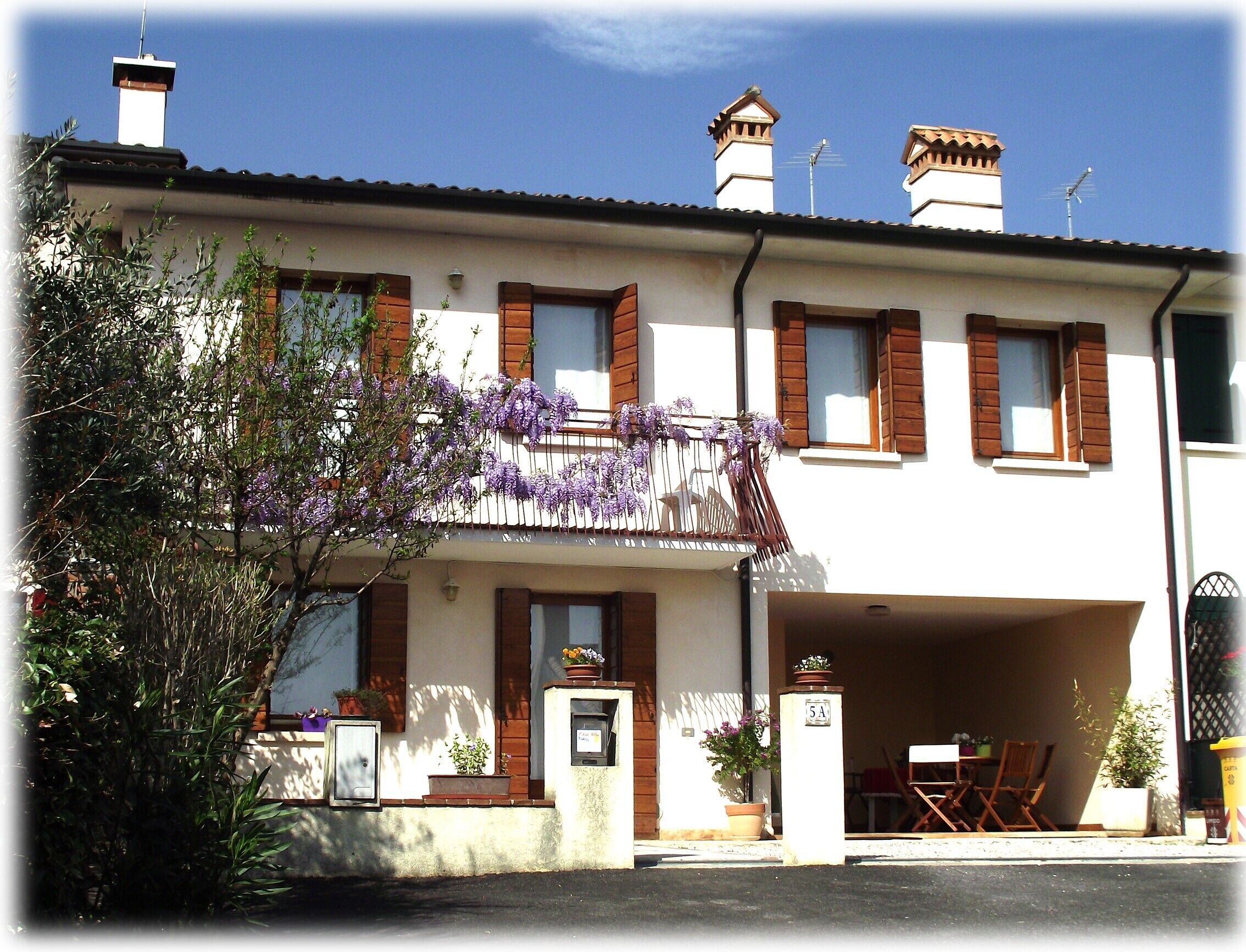 Un bâtiment à deux étages doté d’un balcon orné de fleurs violettes, d’un garage et d’un petit espace extérieur aménagé avec des chaises.