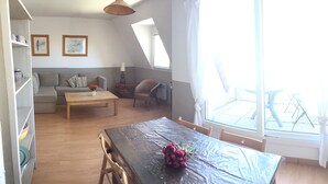 A modern living room with a wooden table, a sofa, a dining table with a plastic cover, and a large window providing a view of the outside.