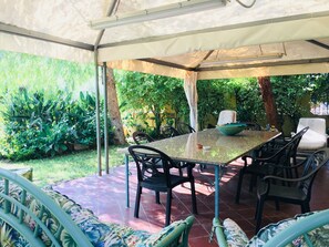 Un patio cubierto con una mesa y sillas, rodeado de exuberante vegetación y piso de baldosas.