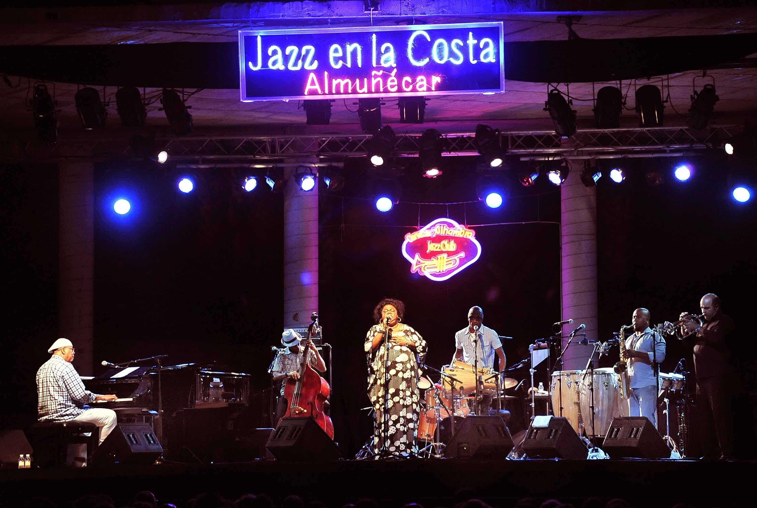 Una banda de jazz actuando en un escenario con un letrero de neón que dice 'Jazz en la Costa Almuñécar'.