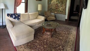 Een woonkamer met een beige bank, een glazen salontafel, een kamerkleed met een patroon, een lamp en een wandtapijt.