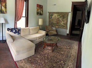 A living room with a beige sofa, a glass coffee table, a patterned rug, a lamp, and a tapestry on the wall.