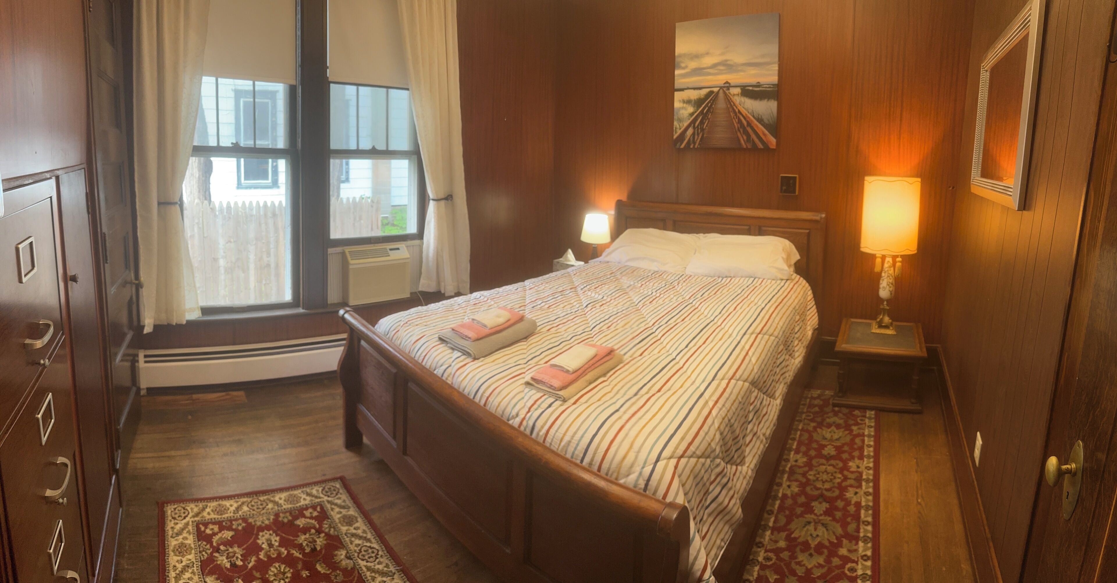 Hotelkamer met een houten bed, een raam met gordijnen, een schilderij aan de wand en een nachtkastje met een lamp.