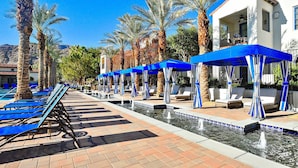 Un espace aménagé au bord de la piscine, avec des chaises longues bleues et des parasols, des palmiers, et un bâtiment doté d’un balcon.