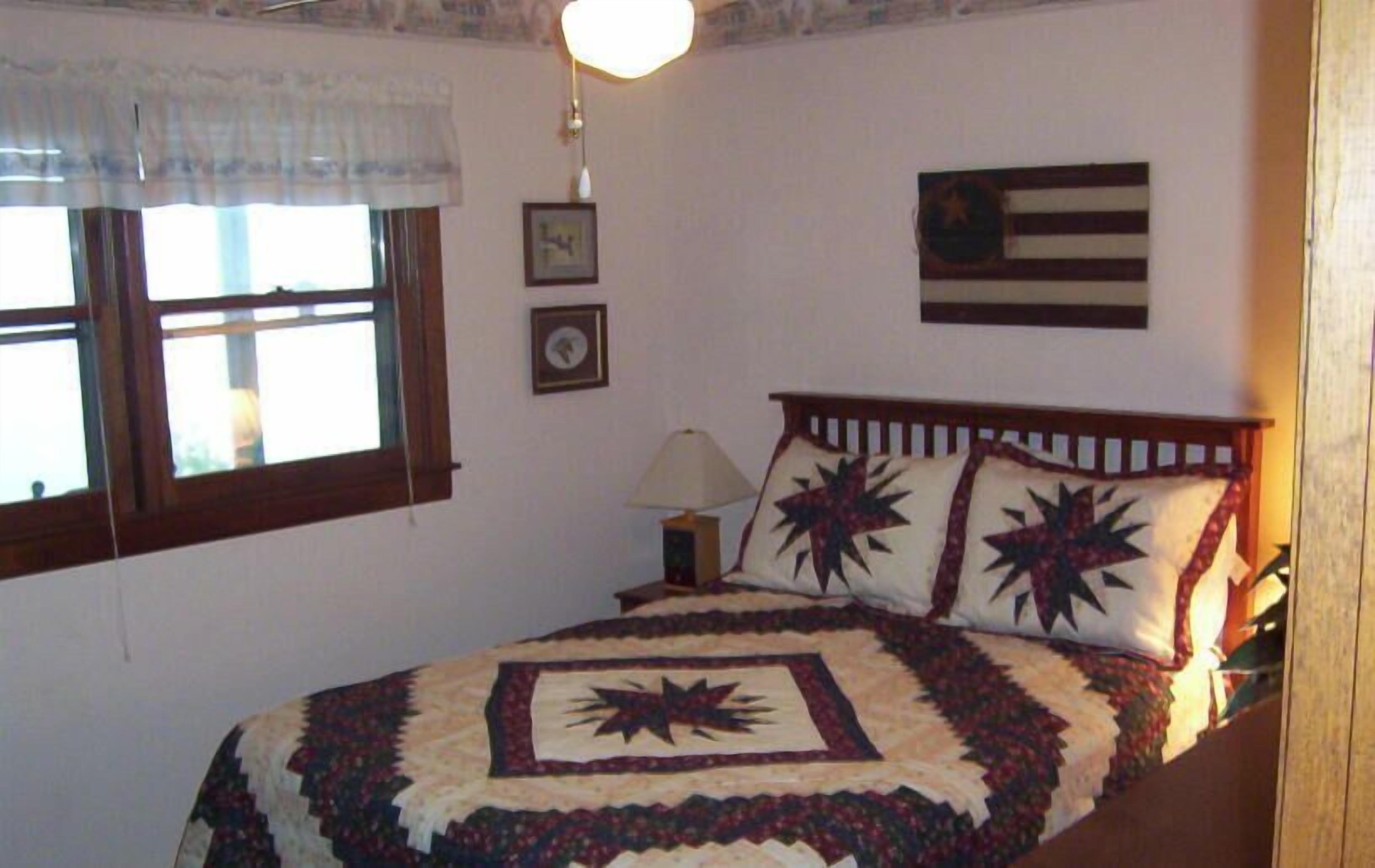 Une chambre à coucher avec un lit, une table de chevet, une lampe, une fenêtre avec des rideaux et un mur orné d’un drapeau.