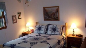 A bedroom with a bed, two bedside tables, a nightstand, a lamp, a framed picture on the wall, and a window with curtains. - A Little Slice of Smokey Mtn. Heaven - with WIFI (Franklin)