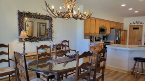 A dining area with a round glass table, wooden chairs, a chandelier with antler motifs, and wooden cabinets in the background. - Snowcreek Resort Vacation Rental 506 (Mammoth Lakes)