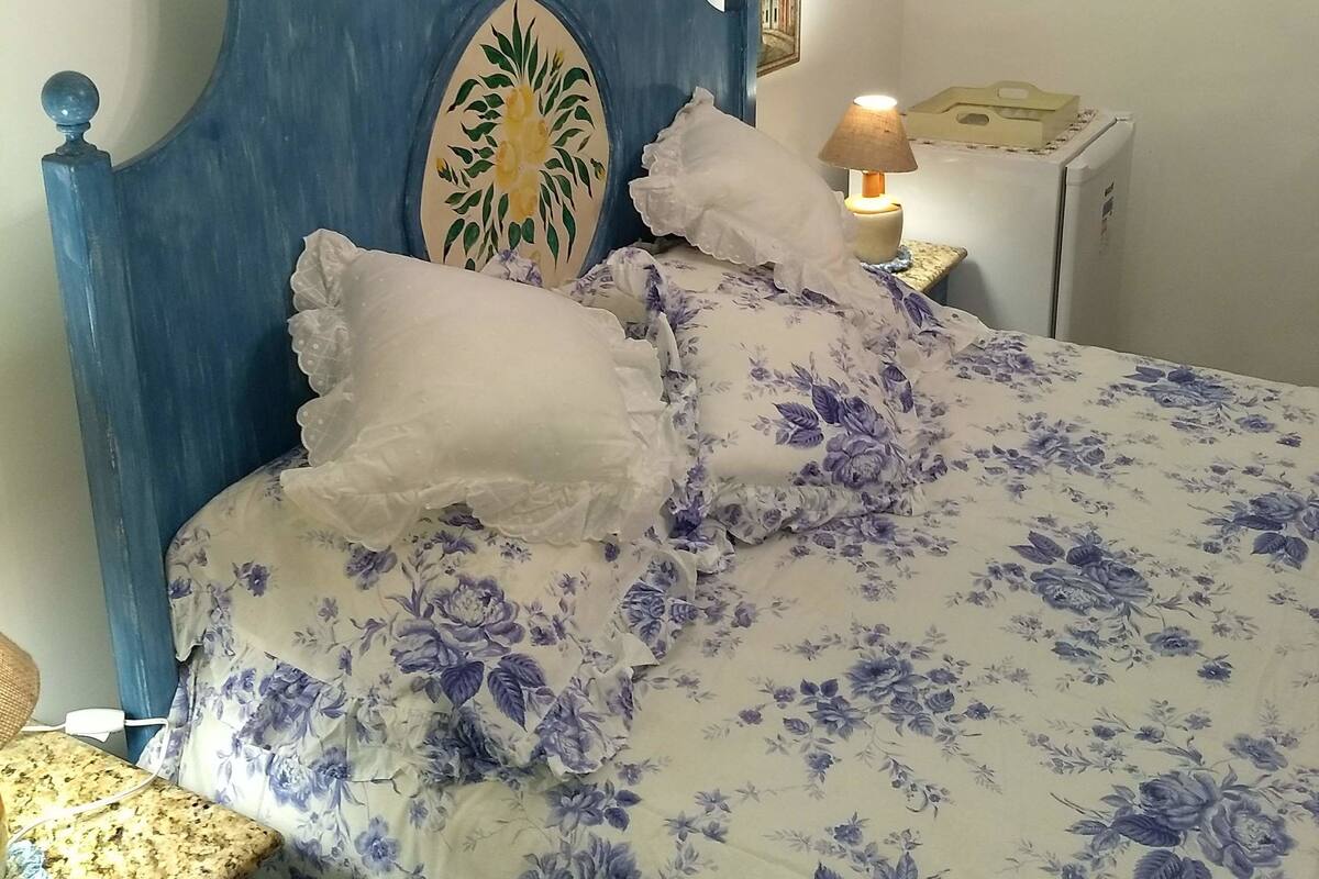 Ett sovrum med en blå sänggavel, ett blommigt sängöverkast, vita kuddar, ett nattduksbord med en lampa och inramade konstverk på väggarna.