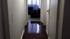 A narrow hallway with wooden flooring leading to a dark-colored sofa and a window with curtains.