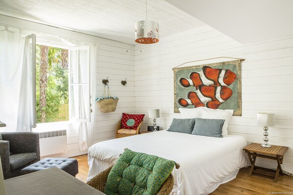 A bedroom with a bed, pillows, a hanging light fixture, a wall-mounted painting, and a window with curtains.