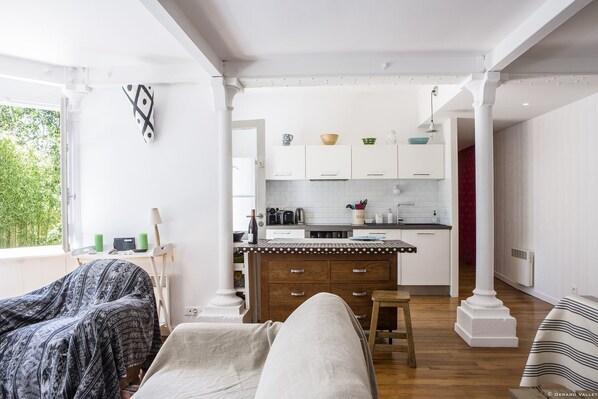 Une pièce confortable avec un lit, une chaise et une vue sur une cuisine dotée de meubles blancs et d’un plancher en bois.