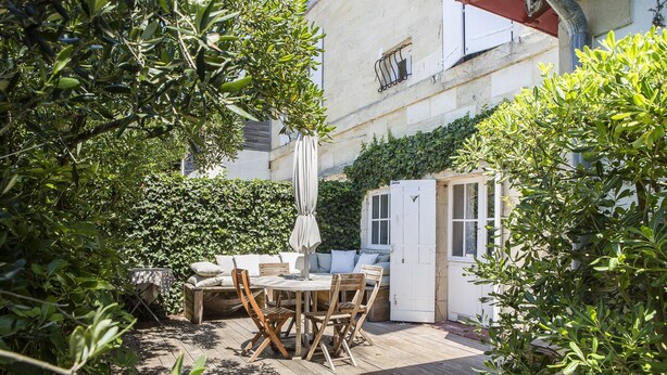 Una terraza de madera con una mesa y sillas, rodeada de follaje y un edificio blanco con ventanas.