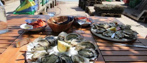 Une table en bois, sur laquelle sont disposées des assiettes d’huîtres et des verres de vin, est placée sur un quai, avec des caisses et une étendue d’eau en arrière-plan.