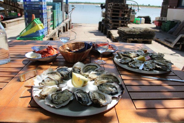 Una mesa de madera con ostras en platos y copas de vino, sobre un muelle con cajas y un cuerpo de agua al fondo.