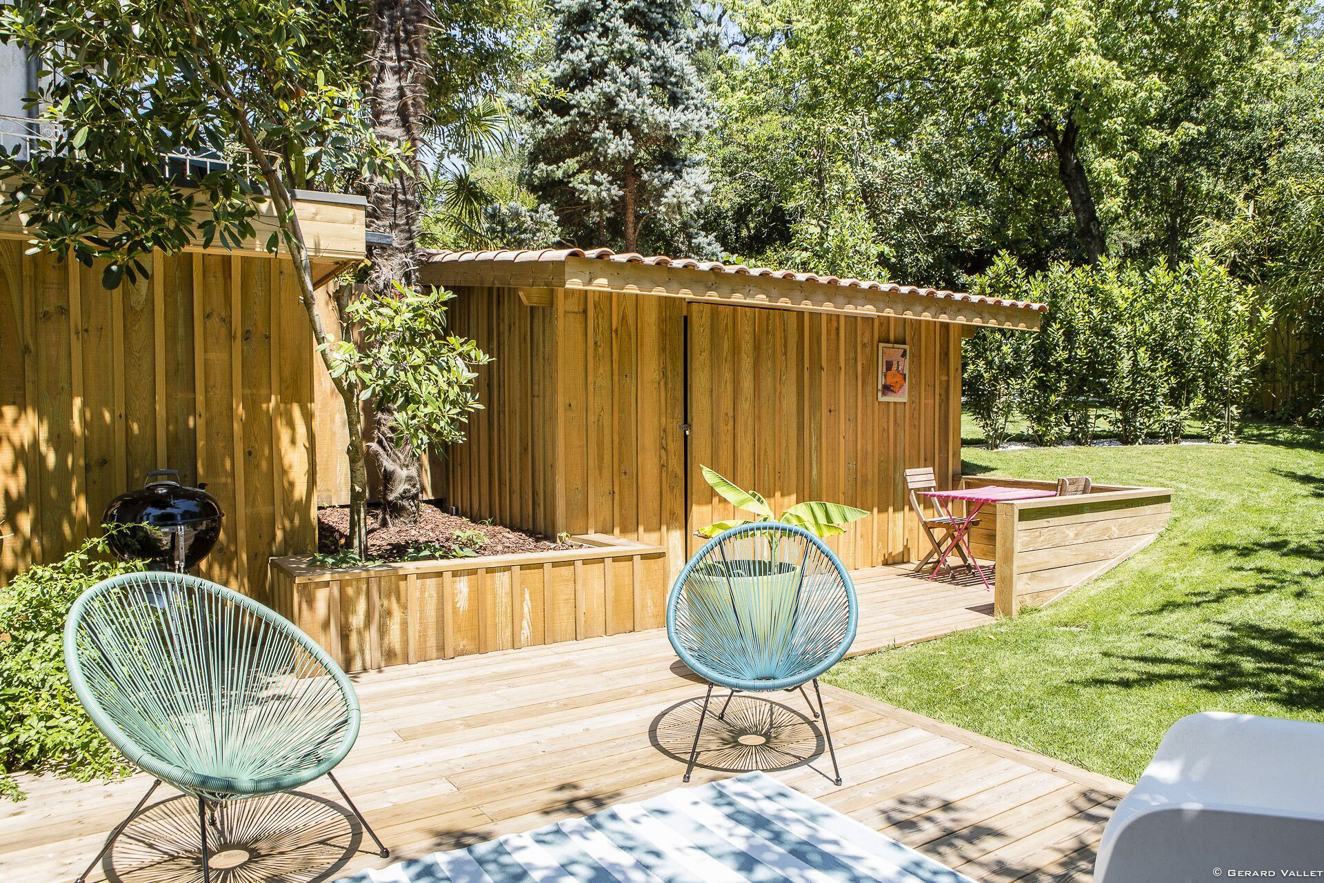 Una terraza de madera con dos sillas de mimbre, un cobertizo de madera pequeño y una parrilla.