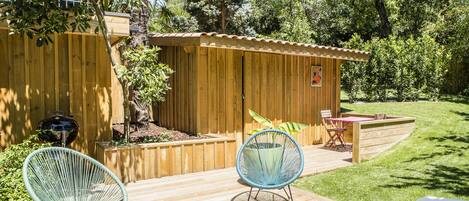Una terraza de madera con dos sillas de mimbre, un cobertizo de madera pequeño y una parrilla.
