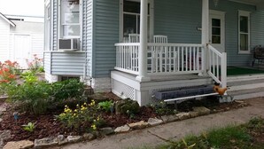 Una casa de dos pisos con un porche cubierto, un pequeño jardín con flores amarillas y un comedero para pájaros.