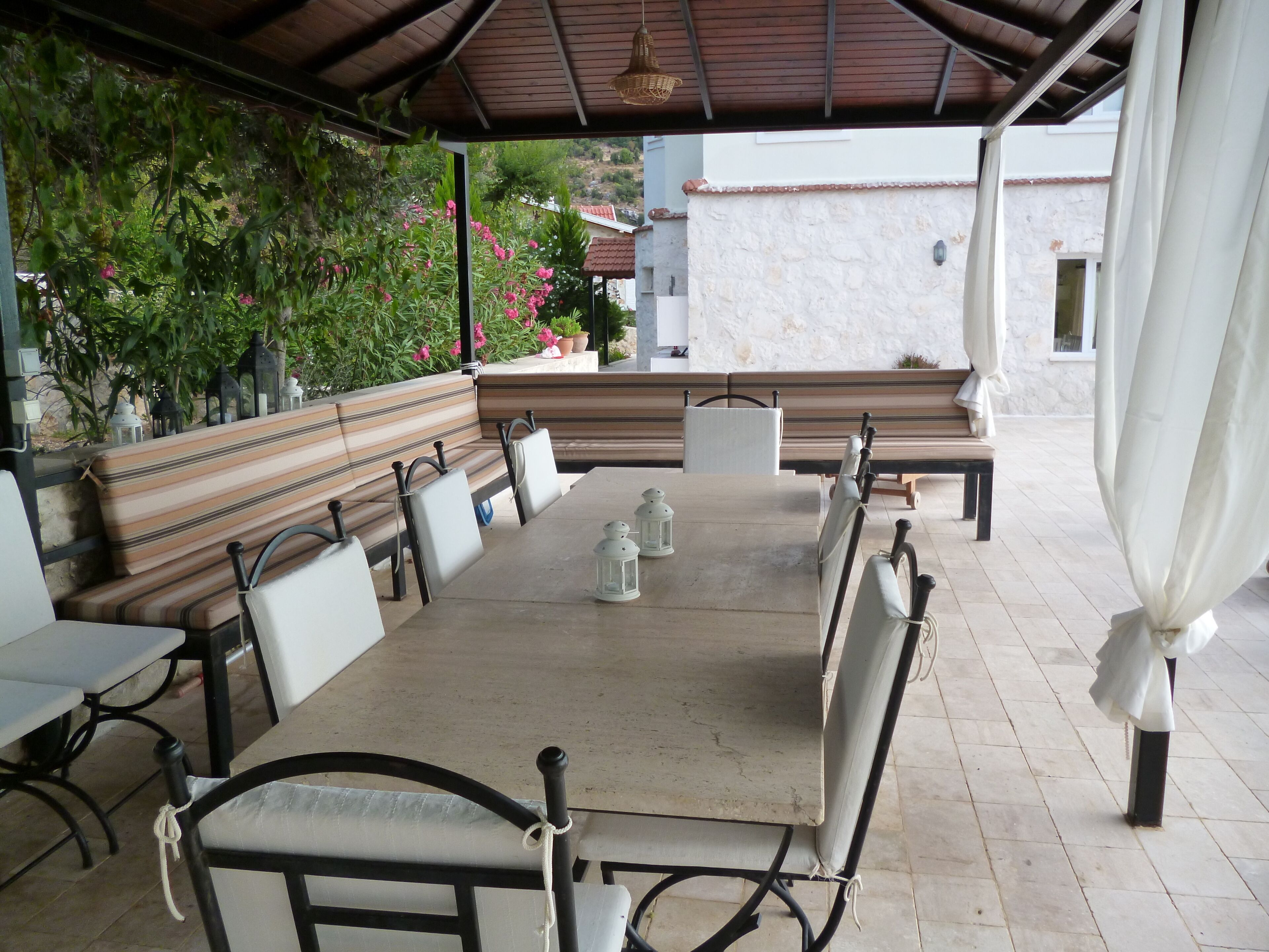 Een terras buiten met een lange tafel en bankjes, omringd door groen en een stenen muur.