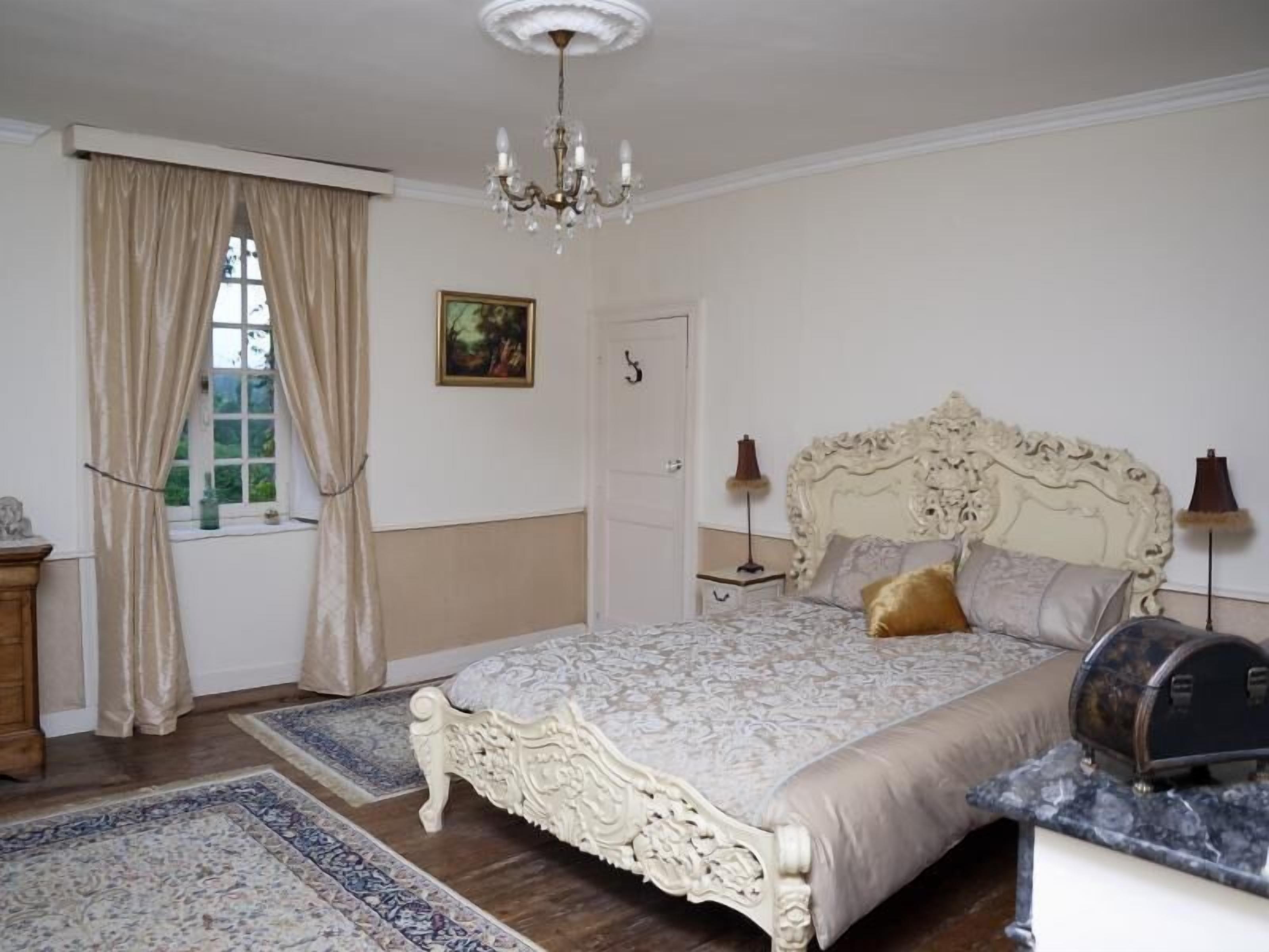 A bedroom with a large bed, a chandelier, a window with curtains, a door, and a small table with a lamp.