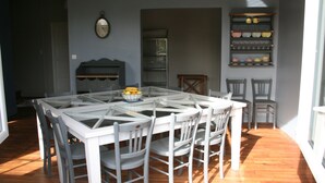 Een eetruimte met een tafel met glazen blad, omringd door witte stoelen, een klok aan de muur en een plank met servies.