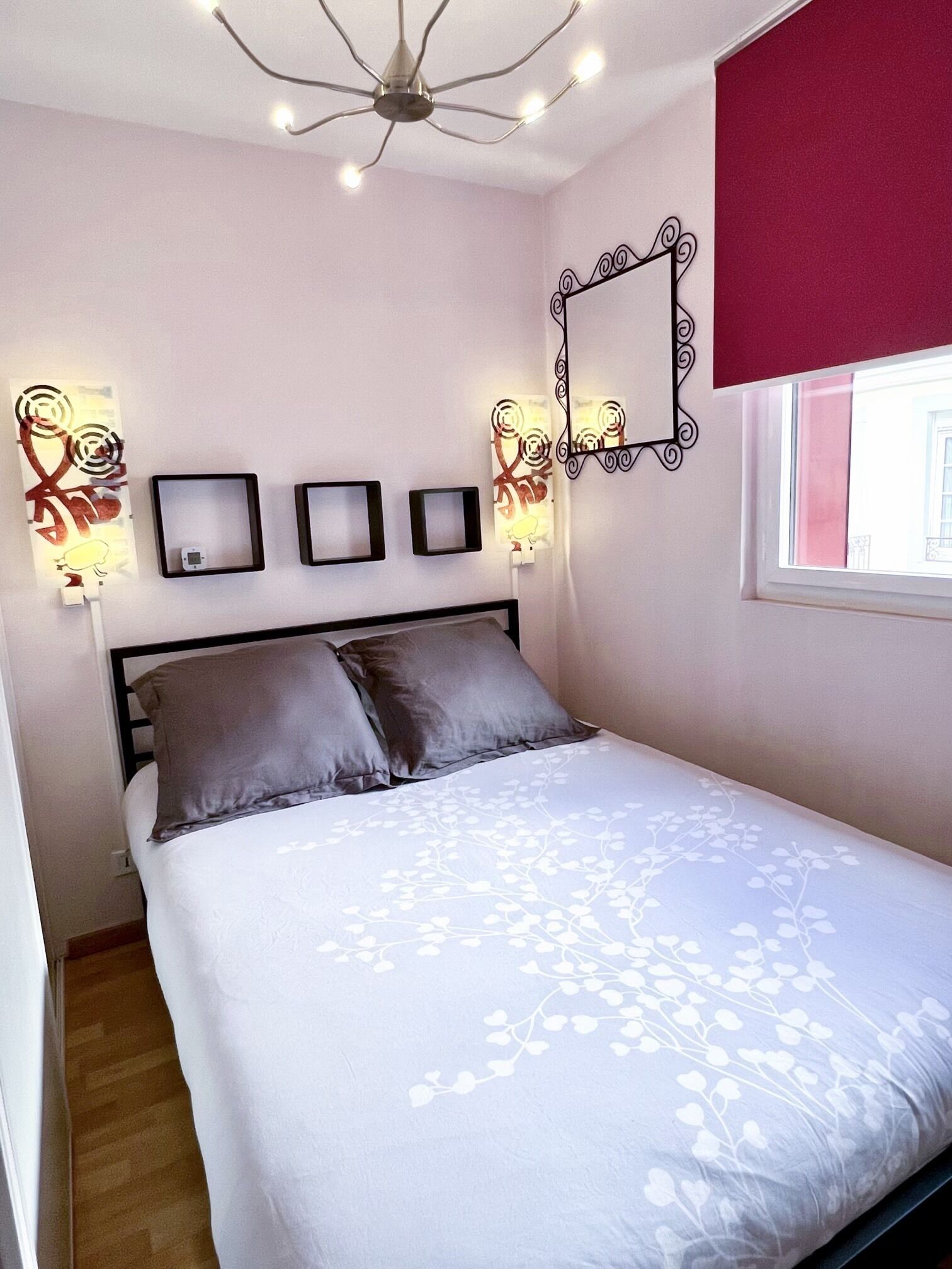 Un dormitorio con una cama, un aplique de techo, lámparas de pared y una ventana con persiana roja.