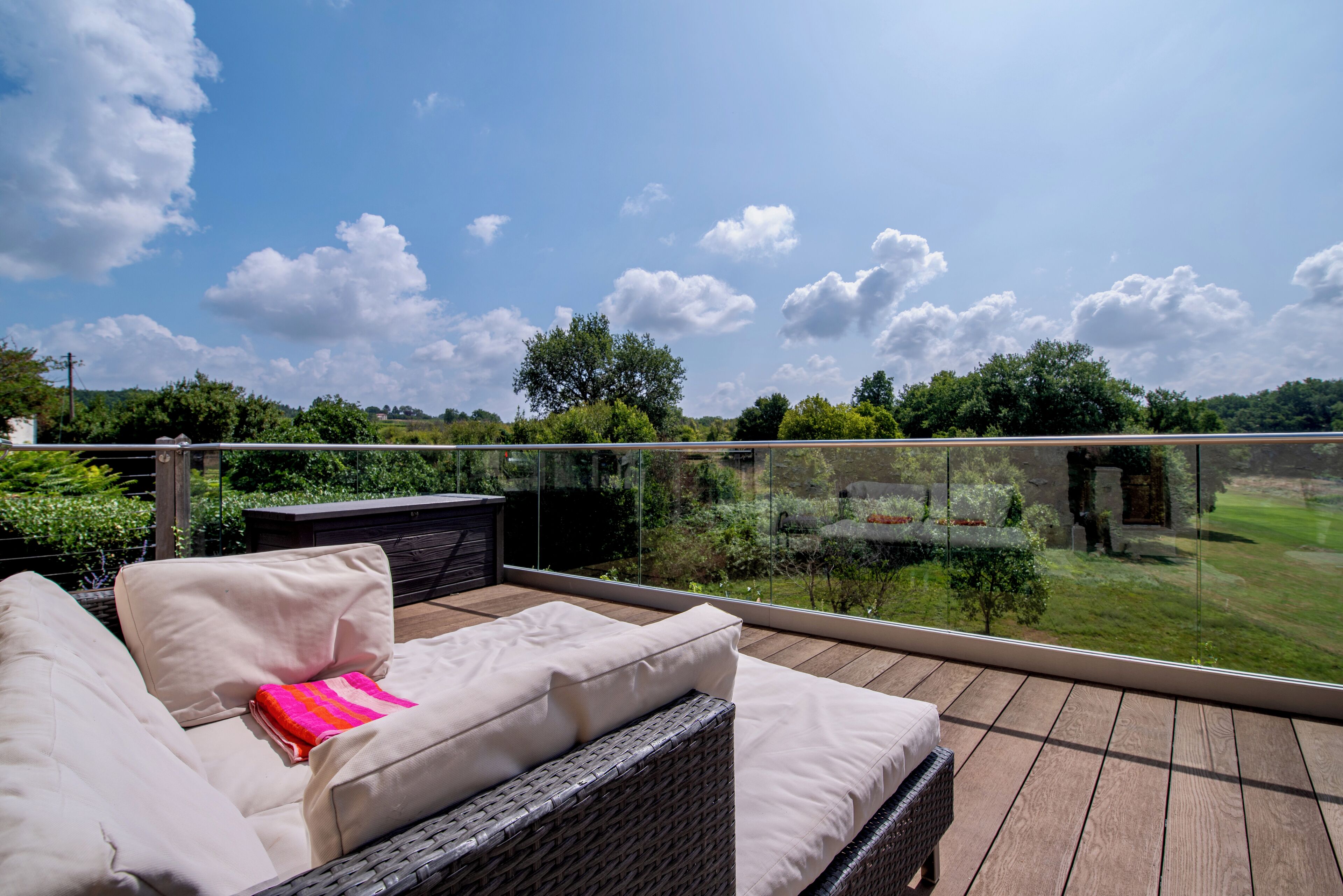 A balcony with a wicker sofa and a small table, overlooking a lush garden and a clear blue sky.