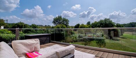 A balcony with a wicker sofa and a small table, overlooking a lush garden and a clear blue sky.