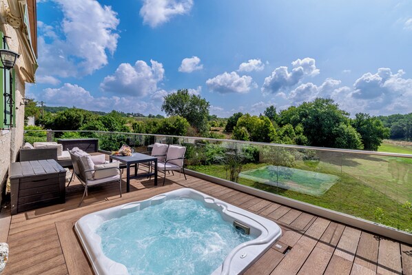 Un jacuzzi en una terraza de madera con mobiliario exterior y vista a un paisaje verde exuberante.