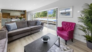 Uma sala de estar moderna com um sofá cinza, uma mesa de centro preta e uma poltrona rosa. Uma grande janela oferece vista para uma piscina e áreas verdes.