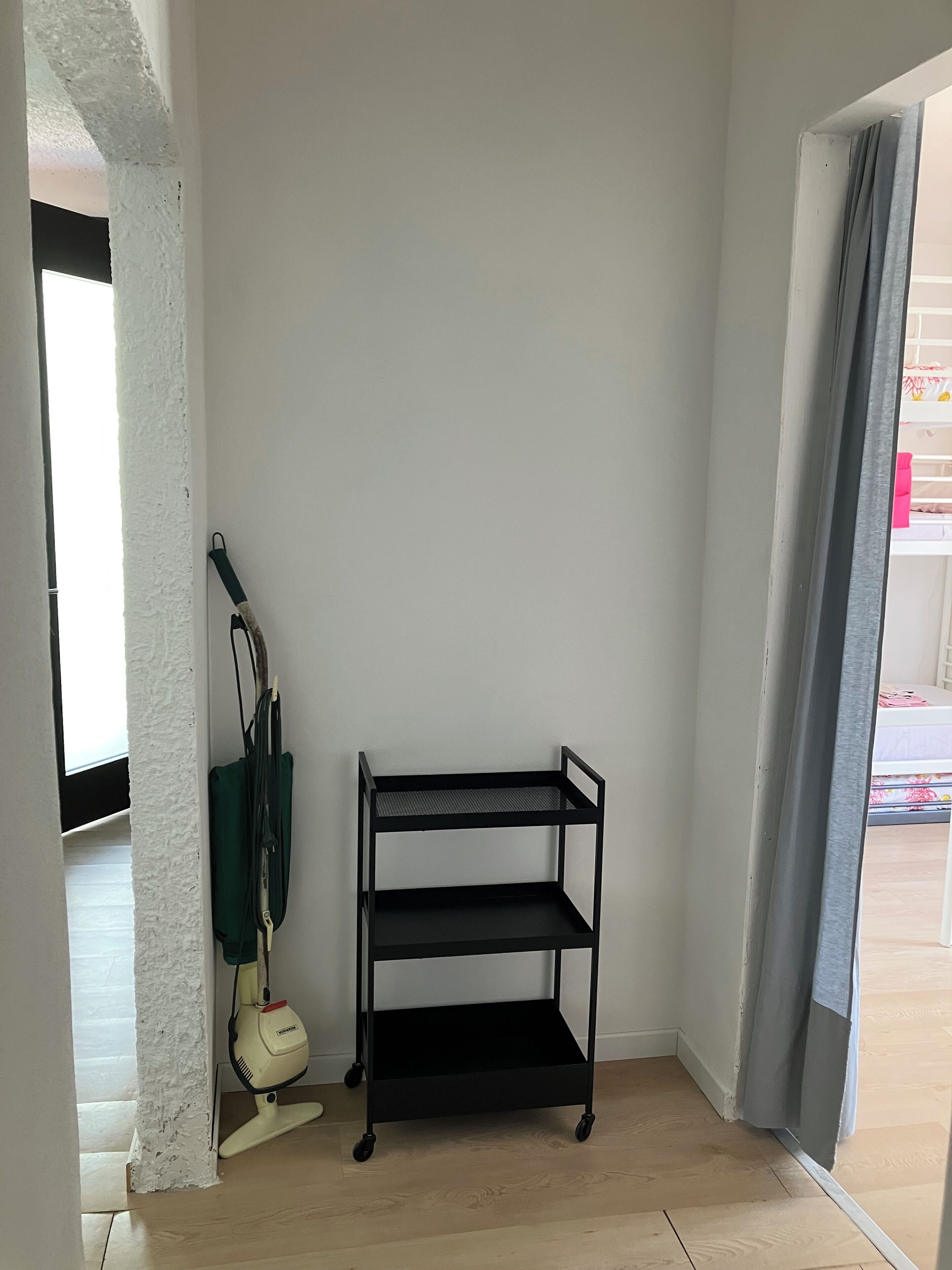 Una habitación en esquina con un estante metálico negro, un aspirador y una pared blanca con textura.