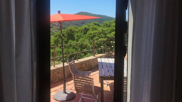 A balcony with a red umbrella, a wooden bench, and a view of a green landscape with trees and a hill.