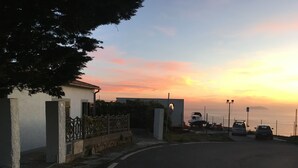 A coastal road with parked cars, a white building with a gate, and a sunset in the background.