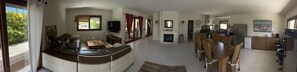A spacious living room with a sofa, a dining table with chairs, a kitchen area with a refrigerator, and a TV mounted on the wall.