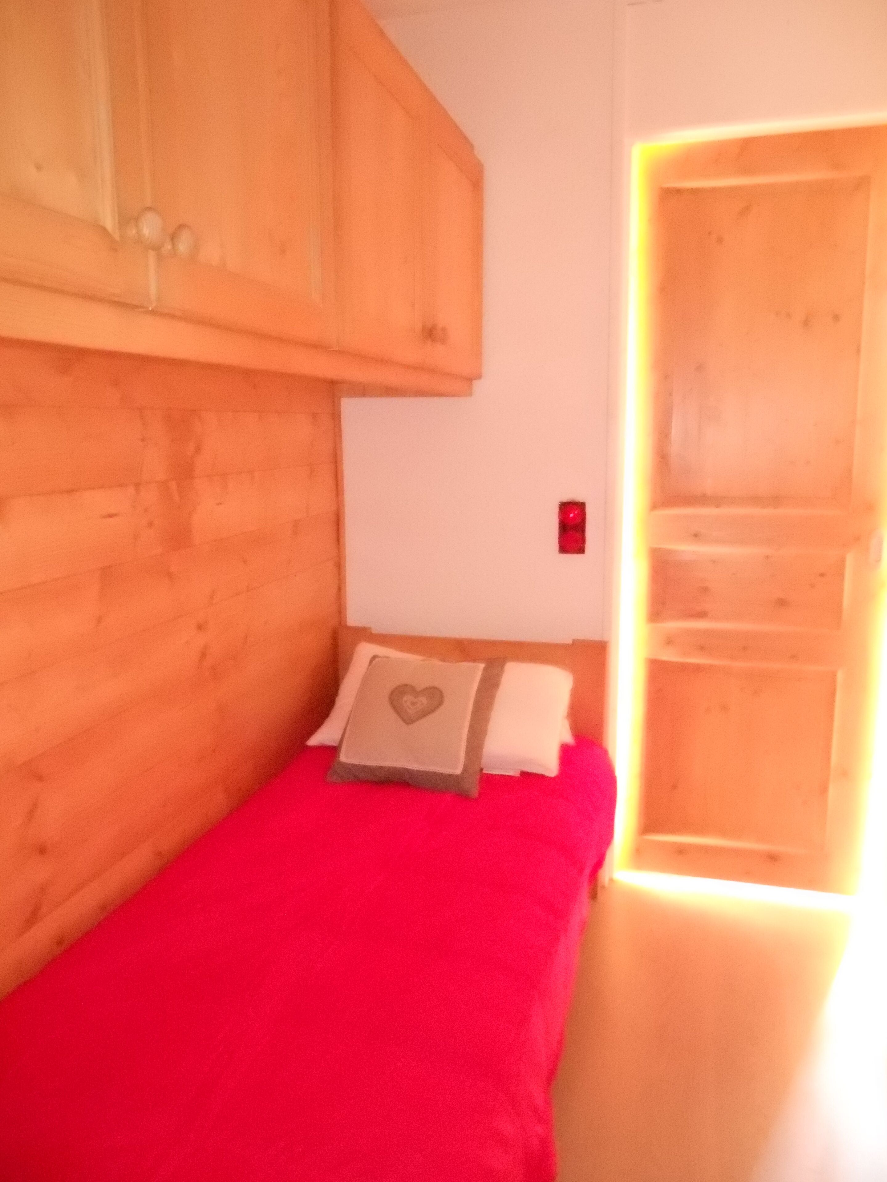 A small, single-bed room with a wooden headboard, a red bedspread, and a pillow with a heart design.