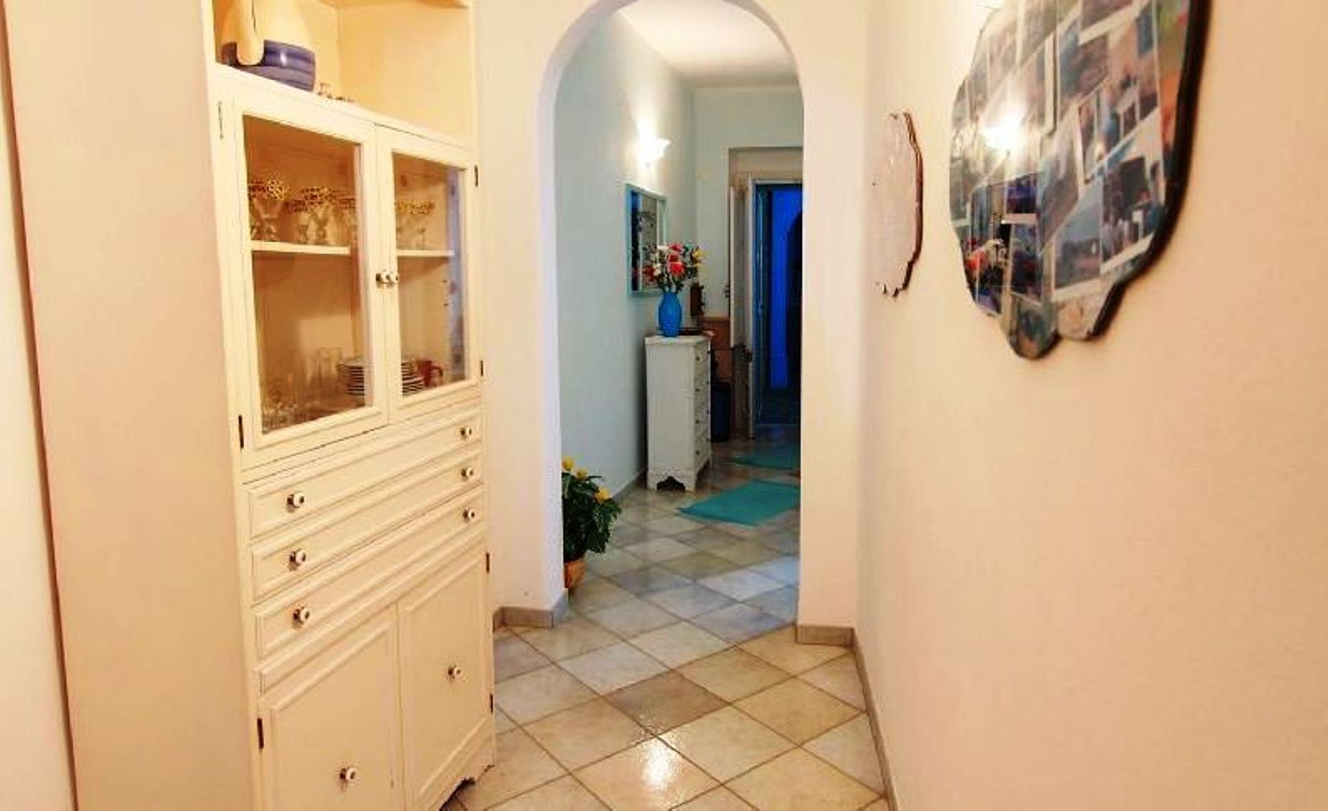 Un pasillo con un armario blanco, una pequeña mesa con un florero y una composición de fotos colgadas en la pared.