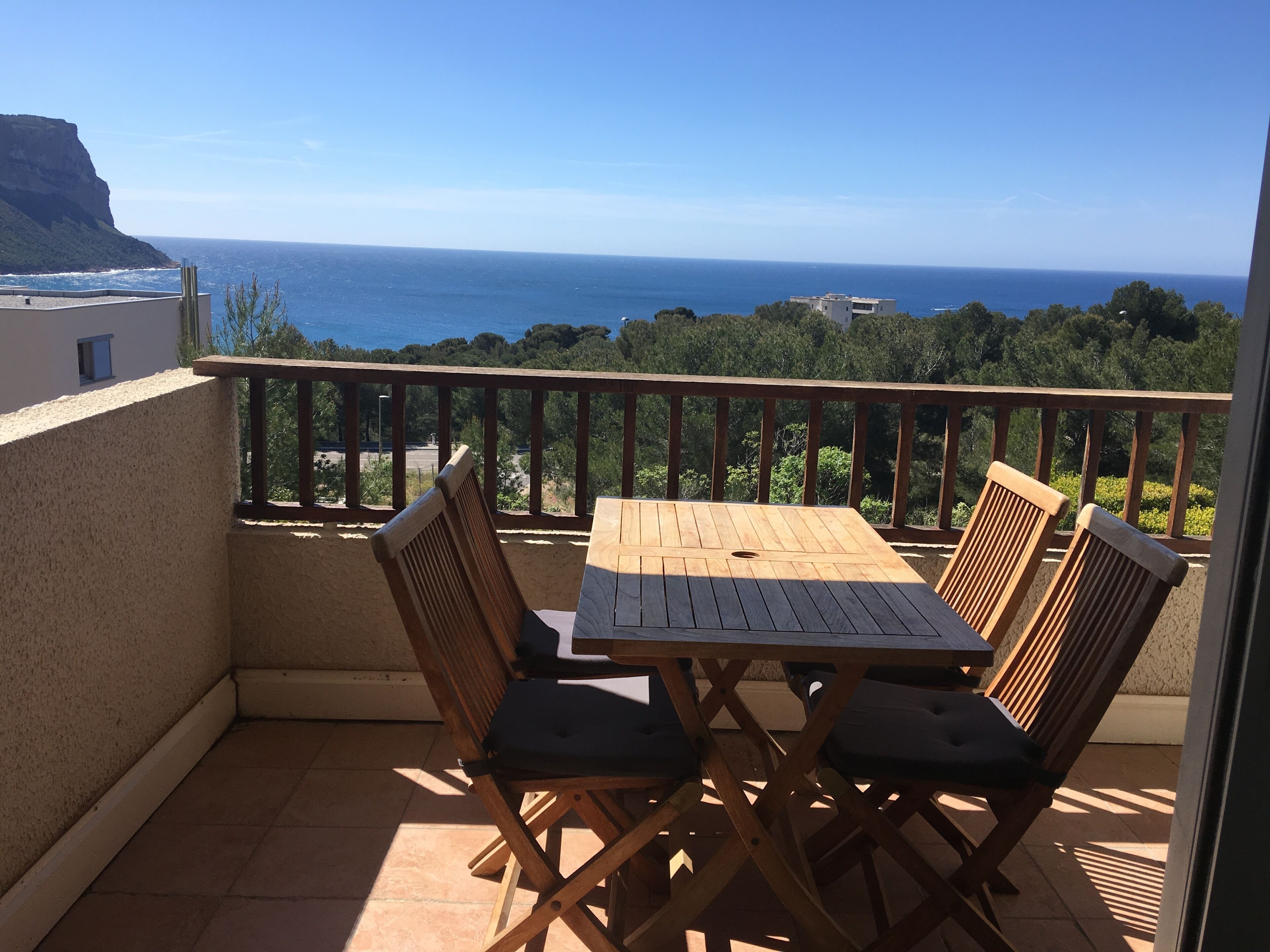 Ein Balkon mit Holztisch und vier Stühlen, der auf eine Küstenlandschaft mit Klippe und Meer blickt.