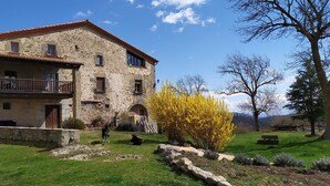 Un edificio de piedra con balcón, rodeado de césped y un arbusto de flores amarillas.
