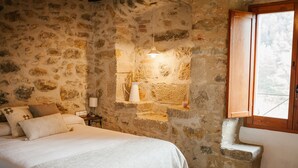 Un dormitorio con pared de piedra, una cama con ropa blanca, una mesita de noche con lámpara y una ventana con contraventanas de madera.