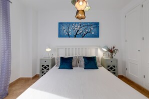 A bedroom with a large bed, two bedside tables, a nightstand, a wall-mounted lamp, a painting of a tree, and a door. - APARTMENT WITH COMMUNITY POOL. 400 METERS FROM THE BEACH. LARGE YARD. (Torrevieja)