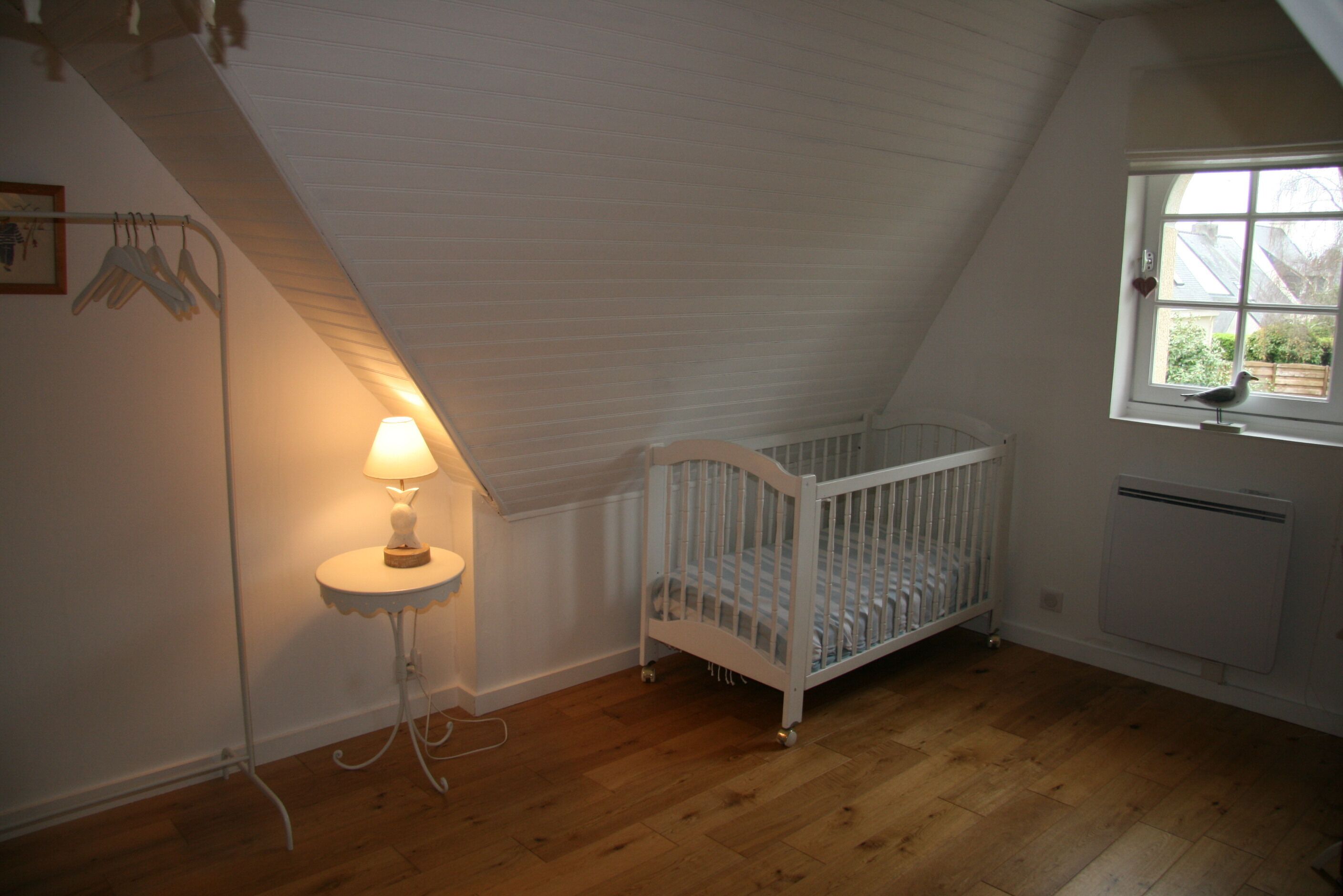 A room with a sloped ceiling, a crib, a small table with a lamp, and a window with a view of greenery.