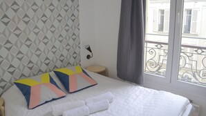 Ein ordentlich gemachtes Bett mit zwei farbenfrohen Kissen, ein Nachttisch, eine Wand mit geometrischem Muster und ein Fenster mit Blick auf Gebäude.